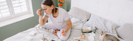 Middle age Caucasian woman applying her makeup in morning. Young woman sitting in bed at home doing her morning routine. Real authentic lifestyle indoor. Web banner header.の写真素材