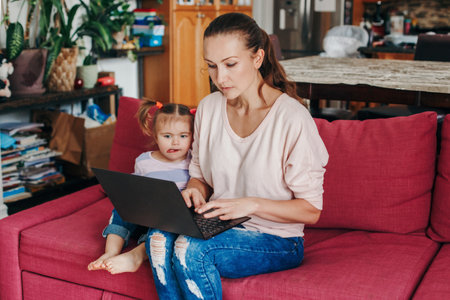 Caucasian mother with baby working on laptop from home. Workplace of freelance business woman with kid toddler. Video chat call. Stay at home single mom earning money at remote job online.の写真素材
