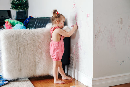 Funny cute baby girl drawing with marker on wall at home. Toddler girl child with milk bottle playing at home. Authentic candid childhood lifestyle moment. Young artist painting on wall in living roomの写真素材