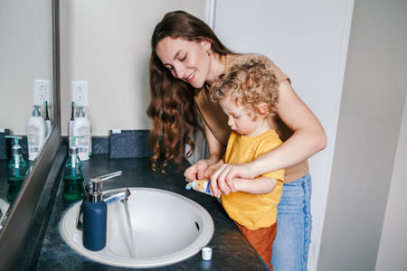 Young Caucasian mother helping boy toddler brush teeth in bathroom at home. Health hygiene and morning routine for children. Mom helping supporting teaching child to use toothbrush.の写真素材