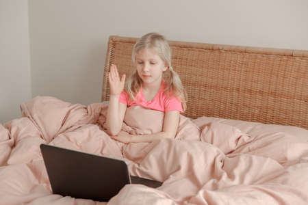 Girl raising hand and learning in virtual online school class. Kid sitting in bed and working on laptop Internet at home. Child using wireless technology education. Homeschooling for children.の写真素材
