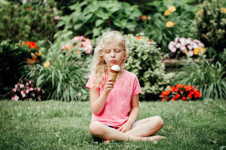 Cute funny adorable girl sitting on grass in park eating licking ice cream from waffle cone. Child eating tasty sweet cold summer food outdoor. Summer frozen meal snack.の写真素材