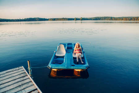 Child girl sitting relaxing in old blue pedal boat tied to wooden dock pier. Beautiful idyllic landscape sunset summer view at Canadian Ontario lake in Muskoka. Water nature place for local vacation.の写真素材