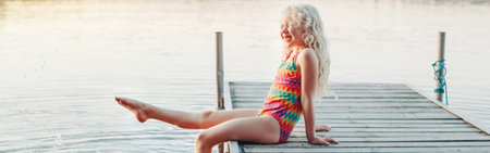 Funny happy cute Caucasian blonde girl child sitting on wooden deck pier by lake. Smiling laughing kid in swimsuit splashing with legs in water. Summer fun outdoor activity. Web banner header.の写真素材