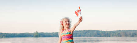 Happy Caucasian girl waving Canadian flag. Smiling child holding Canada flag sitting by water. Kid...