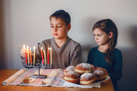 Brother and sister siblings lighting candles on menorah for Jewish Hanukkah holiday at home. Children celebrating Chanukah festival of lights. Dreidel and Sufganiyot donuts in plate on table.の写真素材