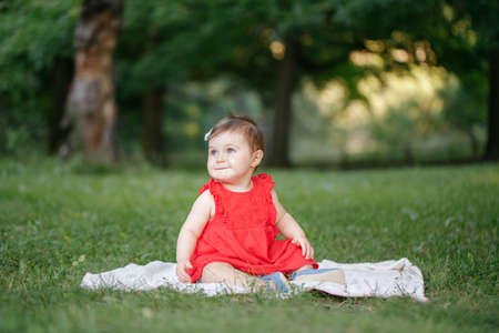 Cute adorable baby girl in red dress sitting on white blanket in park outdoors. Funny child toddler playing and having fun on summer day outside. Funny kid. Authentic lifestyle happy childhood.の写真素材