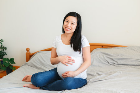 Smiling Asian Chinese pregnant woman sitting on bed touching belly. Expecting young lady in white t-short and blue jeans at home. Healthy happy parenthood lifestyle.の写真素材