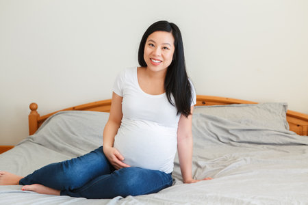 Smiling Asian Chinese pregnant woman sitting on bed touching belly. Expecting young lady in white t-short and blue jeans at home. Healthy happy parenthood lifestyle.の写真素材