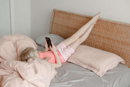 Girl lying in bed at home and chatting with friends on smartphone online. Child kid talking typing on phone in social media. Self isolation on coronavirus covid-19 quarantine. Screen time addiction.の写真素材