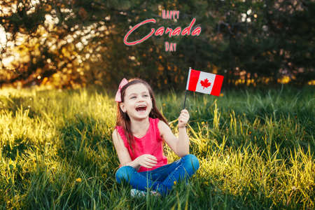 Happy Canada Day. Greeting card with text. Caucasian smiling laughing girl holding Canadian flag. Happy child waving Canada flag in park. Kid citizen celebrating Canada Day holiday outdoors.の写真素材