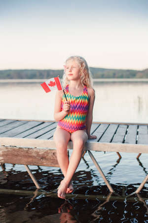 Happy Caucasian girl sitting on dock pier by lake and waving Canadian flag. Smiling child holding Canada flag sitting by water. Kid citizen celebrating Canada Day holiday on first day of July outdoorsの写真素材