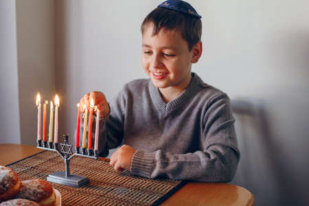 Boy in kippah lighting candles on a menorah for traditional winter Jewish Hanukkah holiday. Child celebrating Chanukah festival of lights at home. Religious Hebrew Judaic Israel culture holiday.の写真素材