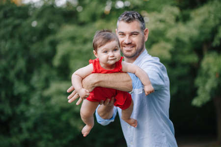 Fathers Day. Middle age Caucasian proud father dad hugging playing with toddler baby girl outdoor. Parent holding carrying lifting child daughter on arms. Authentic lifestyle tender family moment.の写真素材