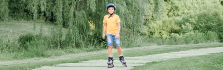 Happy smiling Caucasian preteen boy in helmet riding roller skates on road in park on summer day....