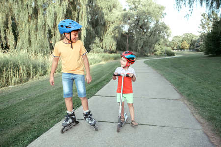Caucasian boys brothers in helmets riding roller skates and scooter on road in park. Seasonal...