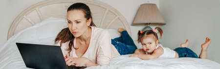 Work from home with kids children. Mother working on laptop in bedroom with child daughter toddler beside her. Funny candid authentic lifestyle family moments. Web banner header.の写真素材