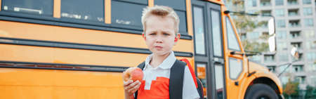 Caucasian boy student with backpack by yellow bus on first September day. Child kid eating apple...