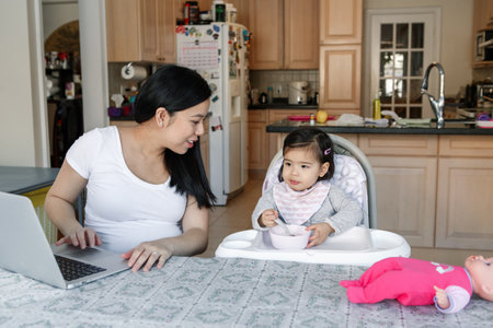 Asian Chinese young mother with daughter baby working online from home on Internet. Workplace of...
