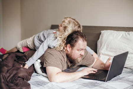 Work from home with kids children. Father working on laptop in bedroom with child daughter on his back.の写真素材