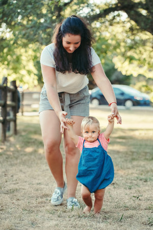 First steps of baby. Cute baby girl learning to walk and holding her mother hand. Mom helping...