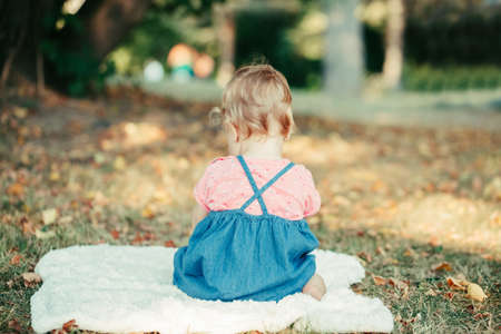 Sad upset baby kid sitting on ground outdoors in park. Child got offended. Upset innocent baby girl does not want to communicate and talk. View from back behind. Cute adorable toddler.の写真素材