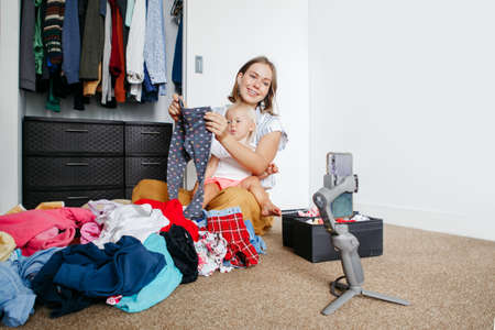Young mother blogger doing video live stream for social media while sorting clothes at home. Video call chat with friends and family on smartphone. Woman using gimbal tripod for video and phone call.の写真素材