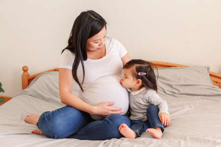 Asian Chinese pregnant woman with toddler girl sitting on bed at home. Girl daughter kid kissing mom...