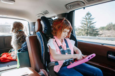Keep kids busy on road trip. Girl sitting in car seat and playing video game on a digital tablet. Kid working on digital pad Internet. Child using wireless technology for education or entertainmentの写真素材