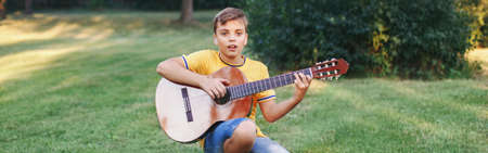 Hard of hearing preteen boy playing guitar outdoor. Child with hearing aids in ears playing music and singing song in park. Hobby art activity for children kids. Web banner header.の写真素材
