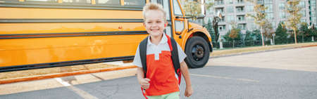 Happy Caucasian boy elementary student running by yellow bus on first September day. Education and back to school in autumn fall. Excited child kid on schoolyard outdoors. Web banner header.の写真素材
