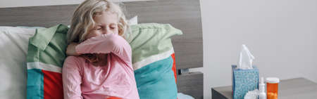 Sick ill kid girl with fever lying in bed at home with flu and sneezing in elbow. Virus cold season flu illness disease. Medicine and health care concept. Web banner or header.の写真素材