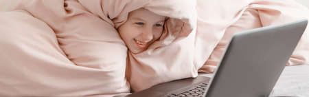Happy girl lying in bed under blanket and learning in virtual online school class. Kid working on laptop Internet at home. Homeschooling education for children. Web banner header.の写真素材