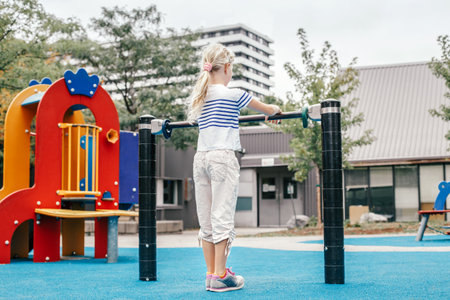 Happy smiling young girl exercising on children pull-ups bar outdoor at playground. Child hanging on high bar. Summer fun activity workout and fitness. Kids sports and recreation. View from back.の写真素材