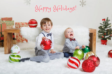 Happy holidays. Christmas card with text. Cute adorable Caucasian boys brothers sitting on white fluffy carpet in festive decorated studio. Traditional Christmas New Year holiday celebration.の写真素材
