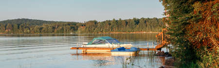 Summer on lake cottage. Small yacht boat by wooden dock pier on lake at sunset. Canadian Ontario Muskoka countryside travel destination. Summer water sport outdoor activity. Web banner headerの写真素材