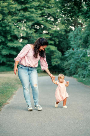 First steps of baby. Cute baby girl learning to walk and holding her mother hand. Mom helping toddler child daughter to go in park outdoors on summer day. Care, assistance and support from parent.の写真素材