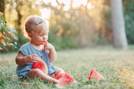Summer seasonal picnic food. Cute Caucasian baby girl eating ripe red watermelon in park. Funny child kid sitting on ground with fresh fruit outdoors. Solid healthy finger food for toddler kids.の写真素材