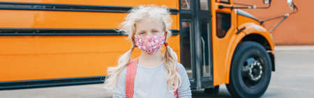 Girl student wearing face mask standing by a yellow bus. Kid with personal protective equipment on...