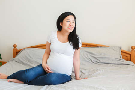 Smiling Asian Chinese pregnant woman sitting on bed touching belly. Expecting young lady in a white plain t-short and blue jeans at home. Healthy happy parenthood lifestyle.の写真素材