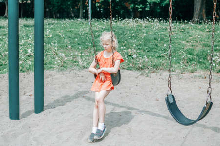 Sad unhappy girl is swinging on swing set in park outdoor alone. Upset bored kid waiting for friend on playground. Lonely child is swinging alone in park and missing friend. Negative emotion.の写真素材