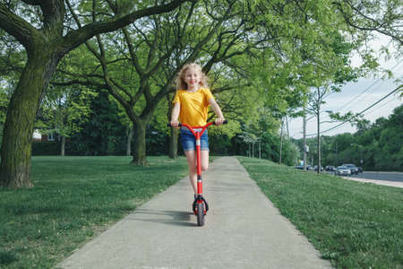 Cute funny Caucasian girl child in yellow t-shirt riding red scooter on street road park outdoor. Summer fun eco friendly sport activity for kids children. Authentic real candid childhood lifestyle.の写真素材