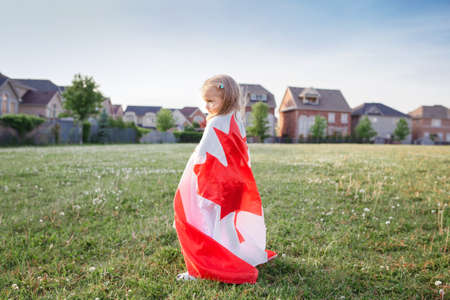 Little girl toddler wrapped in large Canadian flag walking in park meadow outdoor. Canada Day celebration outside. Kid covered with Canadian flag celebrating national Canada Day on 1 of July.の写真素材