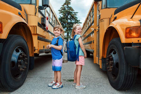 Happy Caucasian children boy and girl kids students standing by yellow school bus. Education and...