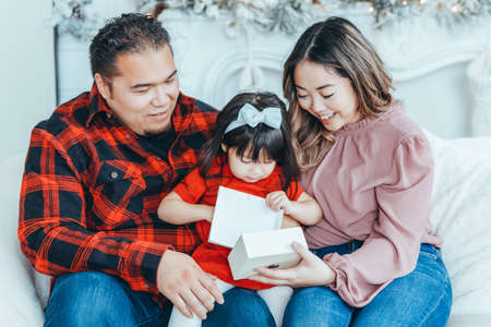 Asian family father, mother giving gift box with present to daughter toddler girl celebrating Christmas or New Year. Mixed race mom, dad, daughter sitting on couch at home. Winter holiday celebration.の写真素材