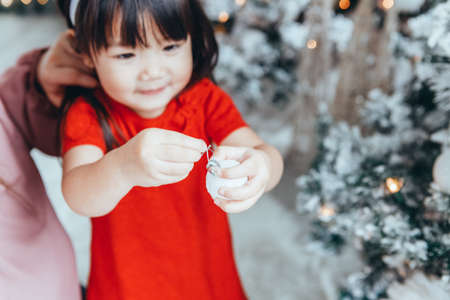 Asian family mother with daughter toddler girl celebrating Christmas or New Year. Mixed race mom, daughter decorating Christmas tree at home. Winter holiday celebration.の写真素材
