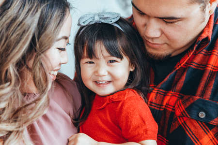 Asian family father, mother with daughter toddler girl celebrating Christmas or New Year. Mixed race mom, dad, daughter decorating Christas tree hugging at home. Winter holiday celebration.の写真素材