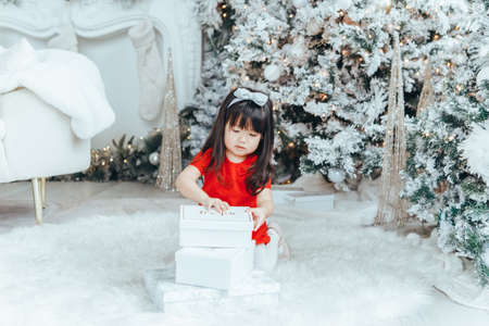 Asian toddler girl in red dress opening gift boxes with presents under Christmas tree at home. Kid child celebrating Christmas or New Year. Winter holiday celebration.の写真素材