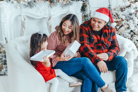 Asian family father, mother giving gift box with present to daughter toddler girl celebrating Christmas or New Year. Mixed race mom, dad, daughter sitting on couch at home. Winter holiday celebration.の写真素材