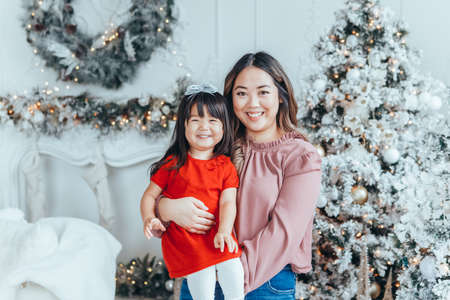 Asian family mother holding daughter toddler girl celebrating Christmas or New Year. Mixed race mom with kid child standing by Christmas tree home. Winter holiday celebration.の写真素材